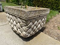 Photo of square concrete planter showing basket weave detail and succulent plants inside, placed on outdoor surface in daylight.
