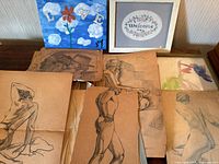 Photo showing multiple sketches and artworks arranged on a table, including nude figure sketches, architectural drawing, whimsical flower painting, and framed cross stitch art.