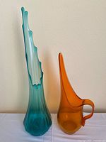 Photo showing both the tall peacock blue Viking art glass vase and the orange swung glass pitcher side by side on a white surface against a beige wall.