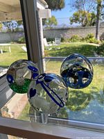 Three glass paperweights displayed with background garden visible through window, showing heart-shaped and two spherical paperweights with internal decorative elements.
