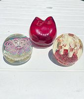 Three items displayed on a white outdoor table under natural light: two Murano glass paperweights and one red punch glass vase.