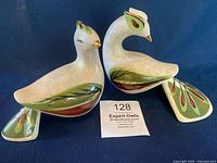 Pair of beige bisque porcelain birds with detailed green, burgundy and gold painted feather and leaf designs, shown from side and front on dark blue fabric background.