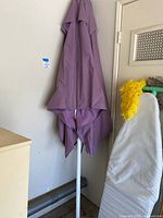 Purple patio umbrella closed and leaning against the wall indoors, showing fabric canopy with stains and the white umbrella frame.