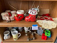 Various Christmas holiday items including ceramic mugs, ceramic candle holder stockings labeled 'Joy', vintage felt Santa with a bell, assorted holiday mugs, antique bells, building block candle holders, vintage ornament, and holiday tin on a wooden shelf.