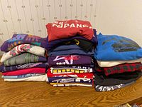 Photo showing three stacks of various sweatshirts and T-shirts, featuring colorful prints and logos.