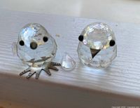 Two small crystal birds on white surface, one missing wing and other missing tail piece visible