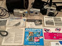 Photo of three Olympus digital cameras with manuals, remote control, and carrying cases spread out on a surface.