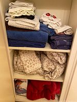 Shelf with stacked towels in various colors and folded beige leopard print flannel sheets and a red lambskin blanket with Sunbeam heating pad box below