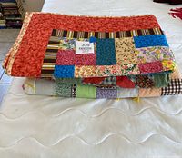 Folded view of 3 colorful handmade quilts stacked, showing various patchwork and fabric designs.