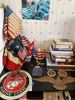 Photo showing vintage American flags, US Marine Corps magnetic seal, stacks of patriotic and historical books, a small eagle figurine, and other small items arranged on a table.