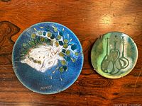 Round enamel metal plate with blue background featuring stylized pendant head, foliage motif, signed Sascha