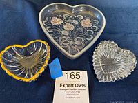 Three vintage heart-shaped trinket boxes shown on a dark fabric background. Left small heart with ribbed clear glass and gold rim, center large heart with pink frosted rose design, right medium clear ribbed glass heart.