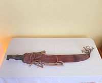 Full length view of machete with leather sheath, laid flat on a white cloth.