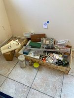 Wide shot of box with assorted vintage sewing supplies including thread, scissors, buttons, small containers, and sewing machine parts on floor.