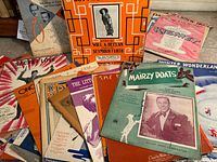 Wide view of 16 vintage sheet music covers showing varied styles and colors with titles and composer information.