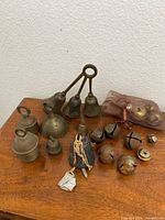 All vintage small brass bells arrayed on wooden surface, showing variety in size, shape, and some wrapped with fabric or plastic packed.