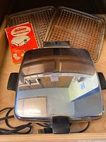Top reflective chrome surface of the vintage Sunbeam waffle baker and grill with two waffle cooking plates in the background and original instruction booklet.