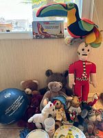 Wide shot of vintage toy group showing plush animals, crash test dummy doll, toucan plush and original box, Sesame Street dish cup, and assorted toys.