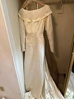 Full length view of vintage wedding dress hanging, showing off-shoulder design, long sleeves, and buttons on sleeve cuffs. Dress fabric has a silky sheen with visible train.