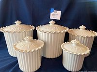 Full view of all five white ceramic canisters showing ribbed bodies and scalloped lids with decorative knob handles, positioned on dark blue fabric background.