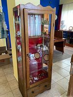 Front and side view of vintage curio cabinet with wood frame and glass panels. Visible scalloped wooden top and bottom drawer with knob, red velvet lining on back panel, and glass shelves inside.