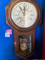 Front view of vintage Ansonia Clock Co. wall clock showing octagonal clock dial with Roman numerals and outer calendar ring, and lower glass window with visible pendulum inside. Dark wood cabinet has visible chips and wear.