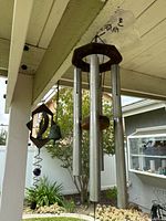Photo of three garden wind chimes hanging from a porch beam with visible yard background. Shows the full hanging lengths and structure of tubes and spiral bell.