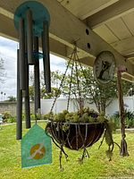 View of silver metal windchime with teal wooden top and bird cutout, macrame plant hanger with succulent plant, and hanging owl thermometer attached to porch beam.