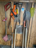 Full view of yard tools hung on wooden wall including trimmer, hedger, pitchfork, shovels, branch trimmer, and gardening tool