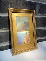 Front angled view of the framed watercolour painting showing gold frame and full artwork of mountainous landscape with sea and foreground foliage.