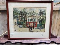 Full view of framed painting showing street scene at Montmartre including cafe and figures walking, with wooden frame and beige mat.