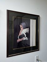 Framed limited edition print by Nancy Noel titled 'Amish Girl with White Cat,' showing the young Amish girl in traditional clothing holding a white cat, side angle view.