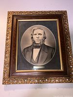 Close-up photo showing detailed view of framed charcoal portrait, ornate carved gold and dark wood frame with glass, oval artwork of man with beard.