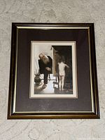 Framed Paul Peel art print showing an elderly man watching a nude child painting at an easel, set in a brown mat and dark wood frame with gold accent