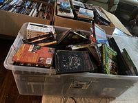 Plastic storage bin filled with various DVD and CD cases showing colorful cover artwork. Titles like Mortal Kombat and Ghostbusters II are partially visible.