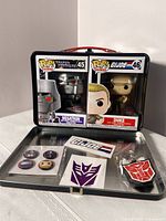 Photo of Funko Pop Megatron and Duke figures inside metal GI Joe lunchbox with collectible pins and patches