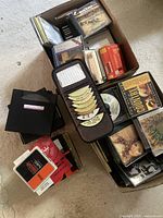 Box filled with CDs and cassette tapes, some visible album covers, multiple plastic cases and cassette cases visible