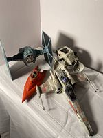 Four Star Wars vehicle toys placed on a white surface including a TIE Fighter, AT-ST Walker, X-Wing Starfighter, and a red Jedi Starfighter.