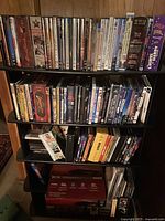 Photo showing multiple shelves holding around 50 DVD and CD cases arranged vertically with visible titles and cover art. Shelving is black. The collection includes a mix of movie and TV show titles mostly from the retro 1990s and 2000 era.
