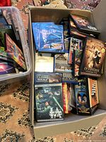 Top-down view of box containing assorted DVDs and CDs with visible titles and cover art for various 1990s and 2000s movies and shows.