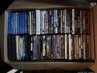 Box of assorted DVDs and CDs, showing spines with titles in a mix of black, blue, and white cases.