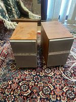 Pair of Panasonic SB-PM18 bookshelf speakers with wood grain finish on a carpeted floor in indoor setting. Speakers are rectangular with gray fabric grille and wood veneer casing.