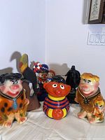 Full view of vintage cookie jar and toy figures including Fred and Barney Flintstone, Ernie cookie jar, Darth Vader figure, toucan, and Americana dolls.