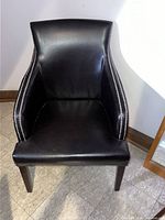 Photo showing a black faux leather accent chair with silver button embroidery along the edges of the armrests and dark wooden legs.