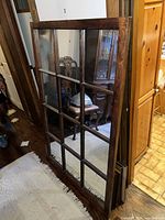 Photo of large rectangular mirror standing against wall, showing wooden frame divided by muntins into 12 mirrored sections resembling window panes.