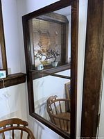 Large rectangular wall mirror with dark wooden frame mounted on a wall, showing reflection of wooden chair and decorative window.