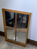 Wood-framed wall mirror with four reflective panes, positioned against a white wall and floor with stone tiles.
