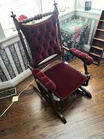 Full view of antique wooden rocking chair featuring red velvet tufted upholstery, carved twisted spindles on arms and backrest, and rockers at the base.
