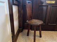 Full view of the carved oak brutalist tripod stool showing its chunky legs, tall backrest with handle cut-out, and the sculptural seat shape.