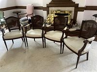 Four Louis XVI style mahogany dining armchairs arranged side by side in a living room setting, showing their overall form, cane backs, light colored cushions, and armrests.